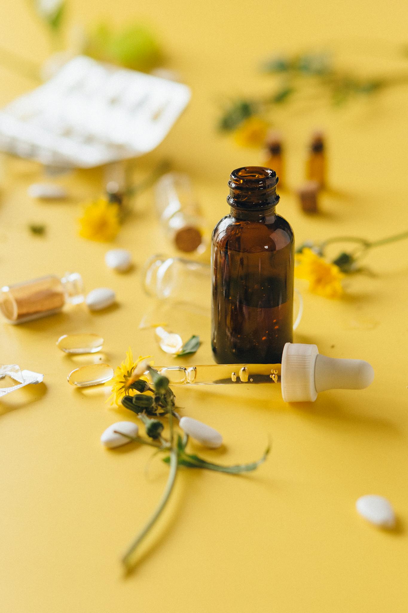 Brown glass dropper bottle among herbs and pills on yellow surface, ideal for wellness themes.